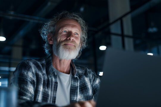 Mature man with gray beard working late on a laptop in a dimly lit modern office, focused and thoughtful while illuminated by cool ambient lights and computer glow.