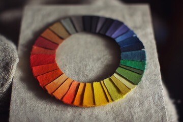 Close-up of a handcrafted color wheel made from felt swatches arranged in a circular rainbow on textured fabric, showcasing vibrant hues, tactile texture and subtle shadow details.