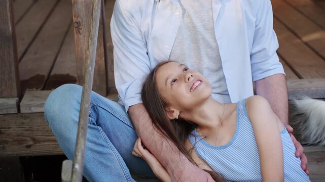 Close-up of daughter lying on father's lap as he sits on porch of wooden country house. Dad hugs, caresses girl, expressing love and care. Single parenting, support, and trust between parent and child