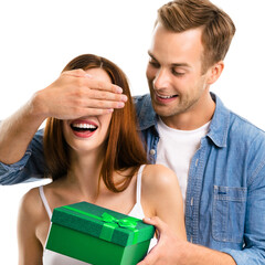 Holiday sales, price discounts, celebrating ad concept - happy smiling man surprises laughing woman by covering her eyes while handing her green gift box. Isolated white background. Square image