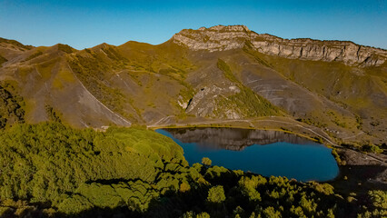 Aerial view of the Caucasus mountains with lush greenery and a serene lake. The landscape features rocky terrain and vibrant vegetation under clear blue skies.