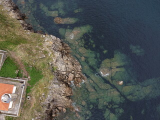 Aerial drone view of Faro de Cee, Galicia, showcasing its red-roofed structure, rocky cliffs, green vegetation, and crystal-clear turquoise waters of the Atlantic Ocean

