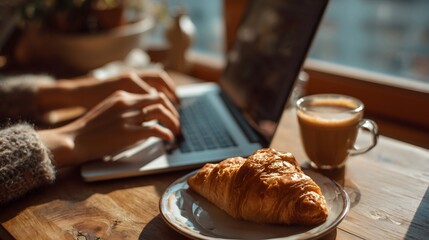 A woman's hands typing on a laptop next to a croissant and coffee cup.