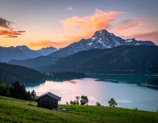 Picturesque alpine lake at sunset. Small wooden cabin sits on grassy meadow at shoreline. Majestic mountain range peaks with snow