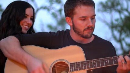 Young caucasian man with neat beard playing acoustic guitar, young caucasian woman with long dark hair singing along by campfire, bokeh background. Cozy romantic evening concept - Powered by Adobe