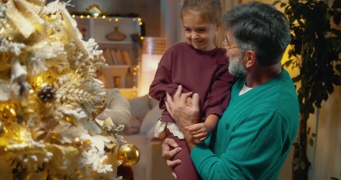 Excited little girl in grandfather&rsquo;s arms placing ornament on Christmas tree at home. Joyful holiday moment of love, family bonding, and festive tradition.