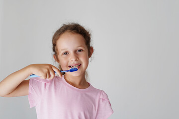 A smiling little girl in a pink t-shirt is brushing her teeth with a blue toothbrush, promoting healthy dental hygiene habits