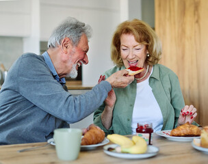 Portrait of an elderly senior couple having breakfast sharing a donut dessert at home. Happy healthy affectionate senior couple eating and sitting at kitchen table having fun enjoying morning meal tog