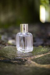A minimalist glass perfume bottle with a silver cap stands on a textured outdoor stone surface, suggesting natural elegance or an organic scent