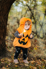 Cheerful child in pumpkin costume standing in autumn forest on Halloween.