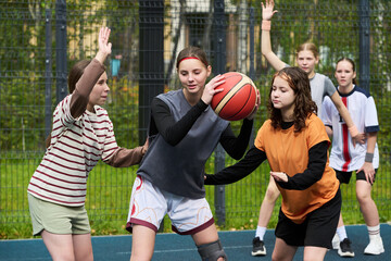 Group of Caucasian teenage girls playing streetball on outdoor court, one girl holding basketball while others defending and raising hands, background showing teammates watching action