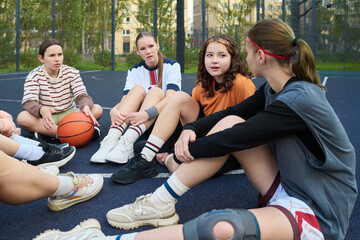 Group of teenage girls sitting on outdoor basketball court resting after streetball game, diverse group including Caucasian and Black teenagers holding basketball, engaging in conversation