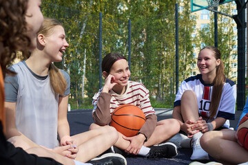 Group of multiethnic teenage girls sitting on outdoor basketball court smiling and talking, one holding basketball, teammates relaxing together after streetball game in sunlight