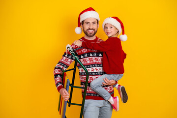 Smiling father and daughter in festive holiday sweaters and Santa hats against a vibrant yellow background, holding a sled