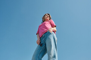 Beautiful carefree young woman with curly hair in pink t-shirt and jeans smiling and looking aside on blue sky background low angle shot.