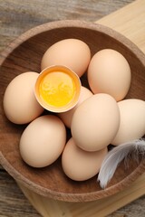Raw chicken eggs in bowl and feather on wooden table, top view
