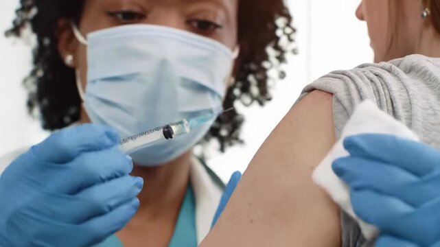 Close-up video of medical professional's gloved hands administering vaccination with chrome syringe on patient's arm in a clean, well-lit setting with bokeh and lens flare. Concept of precise
