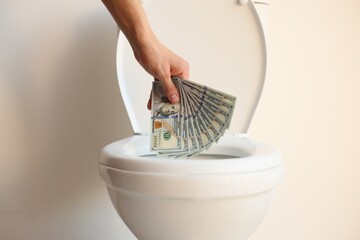 Man throwing dollar banknotes into toilet bowl near white wall, closeup