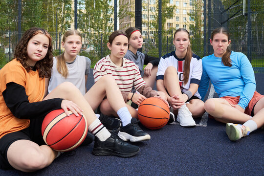 Group of Caucasian teenage girls sitting on outdoor basketball court holding basketballs, looking at camera with serious expressions, urban buildings and trees in background, streetball context