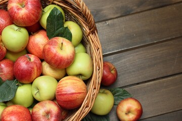 Fresh apples in wicker basket and green leaves on wooden table, top view. Space for text