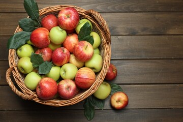 Fresh apples in wicker basket and green leaves on wooden table, top view. Space for text