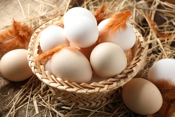 Raw chicken eggs in wicker basket and feathers on straw, closeup