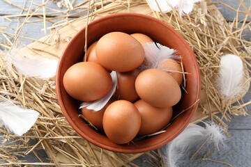 Raw chicken eggs in bowl, feathers and straw on grey wooden table, flat lay