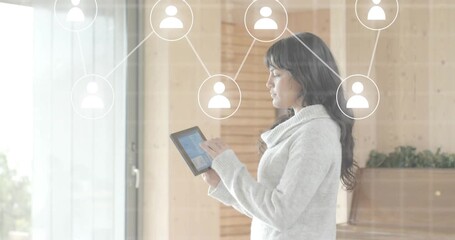 Woman holding tablet by window tapping and swiping analyzing social network connections with icons - Powered by Adobe