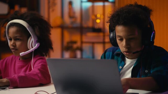Focused children in headsets playing multiplayer game on laptops, teamwork hobby