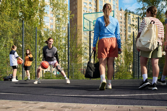 Group of multiethnic teenagers playing streetball on outdoor court, Caucasian teenage boy dribbling basketball while other teenagers standing and watching, urban buildings in background - Powered by Adobe