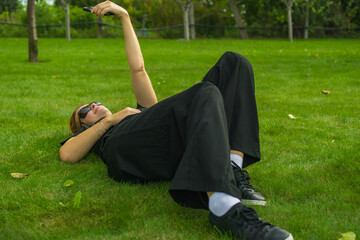 Stylish woman in black clothes and sunglasses lies on a sunny park lawn holding her smartphone above to take a selfie outdoors.