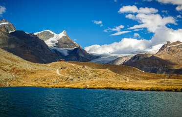A house near a lake on a high mountain slope against the backdrop of snow-capped peaks and glaciers