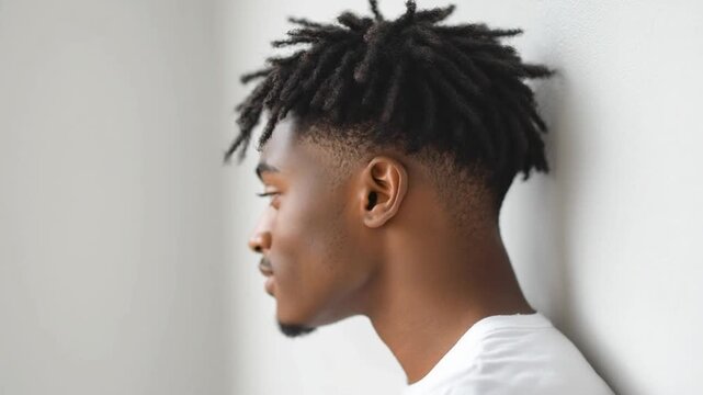 Stylish young man with dreadlocks showcases modern hairstyle against light wall, highlighting texture and shape while exuding relaxed and confident vibe in his profile