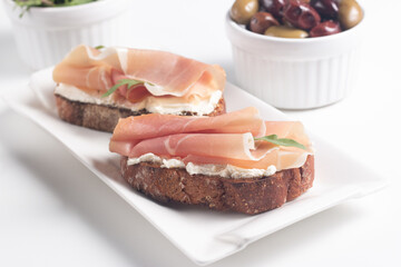 Two bruschettas with prosciutto, ricotta cheese and rucola on ciabatta bread. Olive and spices  decorated.