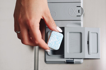 Hand putting dishwasher tablet into dispenser. Cleaning and hygiene concept. Household chore before washing dishes. Modern detergent pod with blue specks for sparkling clean results.
