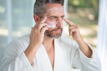 Fototapeta premium Middle-aged man applying facial cream during a morning skincare routine at home. Mature man using a moisturizing mask for anti-aging and rejuvenation. Male cosmetic facial treatment.