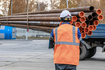 A construction worker in safety gear manages a load of pipes at an industrial site. The setting is...