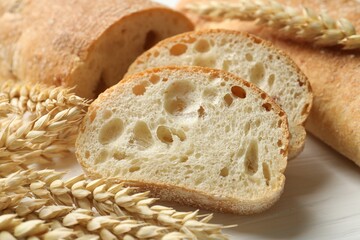 Fresh crispy ciabattas and wheat ears on white wooden table, closeup