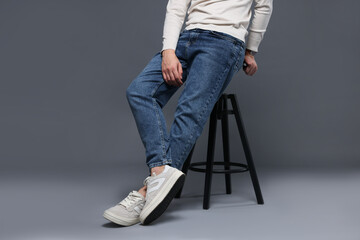 Man in stylish jeans on stool against grey background, closeup