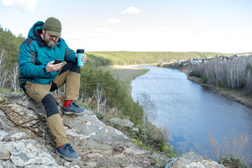 A man is sitting on a rock overlooking a river with a cup of coffee in his hand