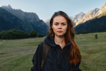 A focused young woman stands outdoors in a rugged landscape. She wears a dark jacket and backpack,...