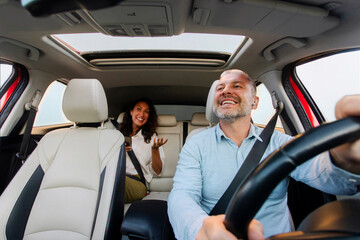 Happy European male driver riding car, looking at lady sitting in auto on back passenger seat and talking, windshield view