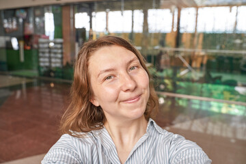 Fototapeta premium Smiling young caucasian female in striped shirt taking selfie in bright indoor setting.