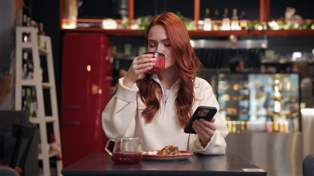 Focused red-haired woman sitting in cozy cafe while sipping drink from ceramic mug. Holding smartphone with one hand. Reading messages or checking updates online while enjoying peaceful moment alone.
