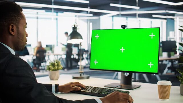 Young african american businessman in dark suit intently typing at sleek desk with bright green screen and tracking markers. High-tech corporate office, visual effects production.