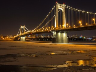 Fototapeta premium A beautifully lit suspension bridge spans a frozen river at night, with city lights in the distance and a dark, starry sky above.