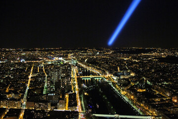 Aerial view of Paris, France, at night, with illuminated streets, bridges, and the Seine River. A bright blue spotlight illuminates the dark sky. Conceptual images for cityscapes, travel, tourism.