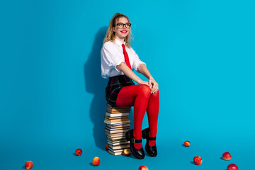 Cheerful young woman sitting on books in colorful outfit and glasses against a vibrant blue...