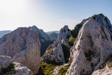A red hiking trail marker is visible on a large boulder, part of the stunning karst scenery of the Velebit mountains in Croatia, illuminated by the bright sun overlooking distant mountain ranges.