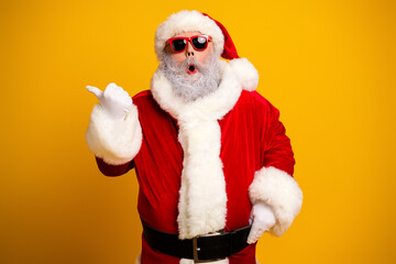 Cheerful Santa Claus in red costume wearing sunglass pointing with excitement against bright yellow background